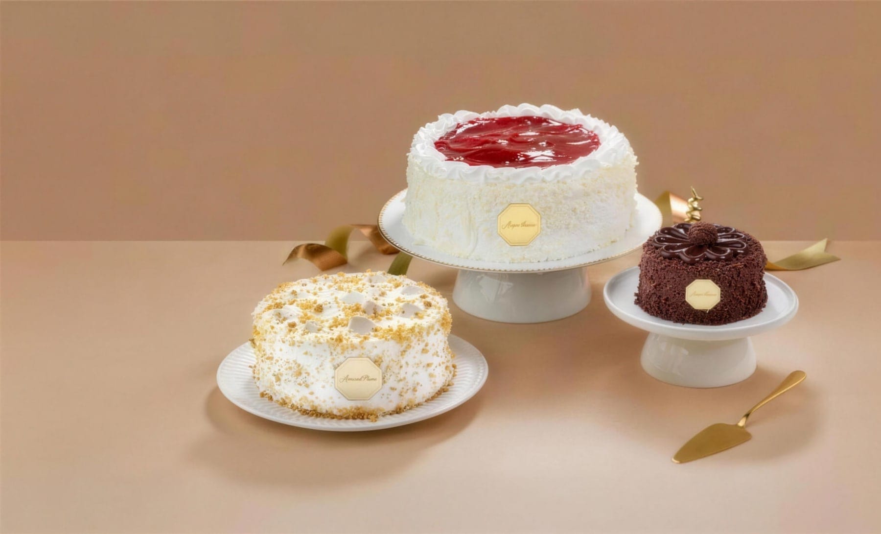 Três bolos Amantícia em suportes de porcelana, com a placa dourada da marca, sobre fundo caramelo